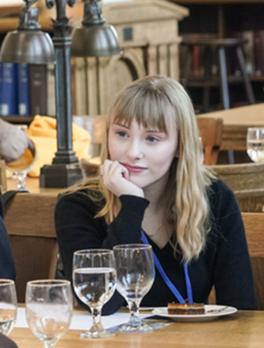 A student listens while sitting at a table with wine glasses and dessert in front of her